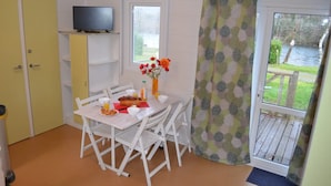 Een kleine kamer met een eettafel, stoelen, een televisie op een plank, een raam met uitzicht op groen en een deur.
