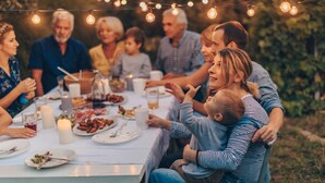 Un groupe de personnes est assis autour d’une table, où sont disposés de la nourriture et des boissons. La scène se déroule à l’extérieur, et des guirlandes lumineuses sont suspendues au-dessus d’eux.