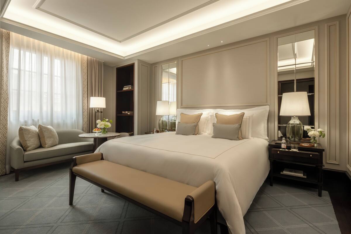 A spacious hotel room with a large bed, a sofa, a bench, a bedside table with a lamp, and a mirror.