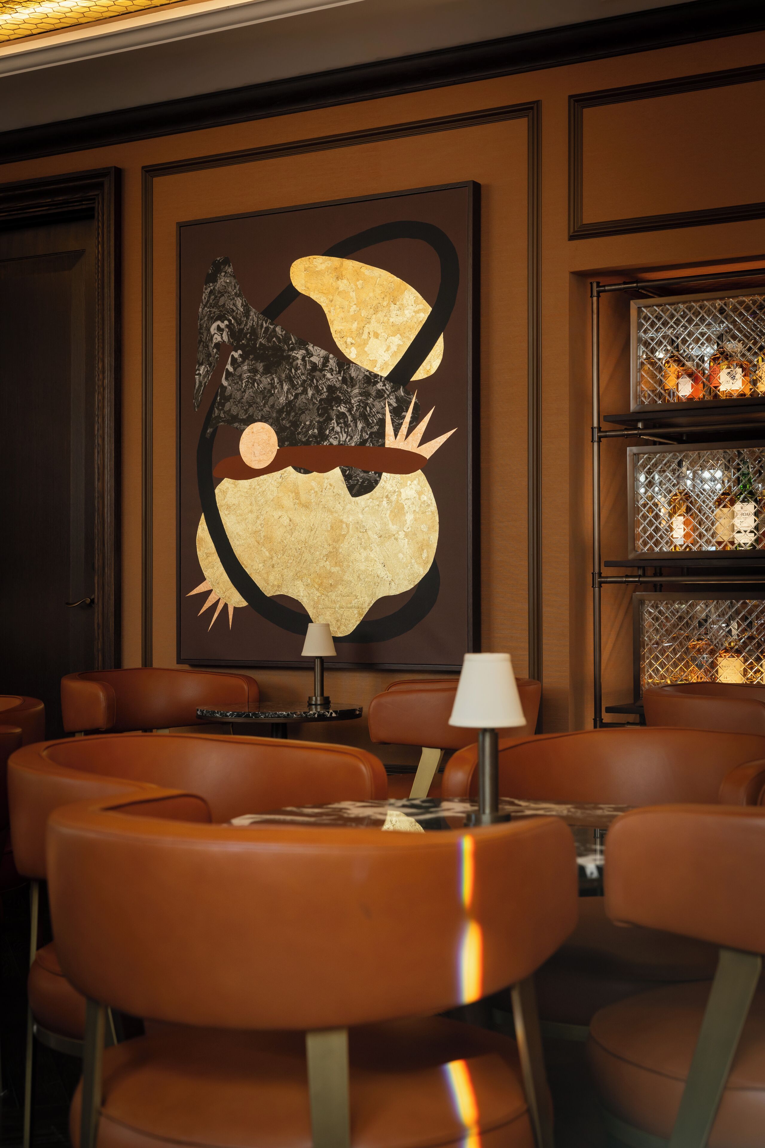 A restaurant with brown leather chairs, a table with a lamp, and a large abstract painting on the wall.