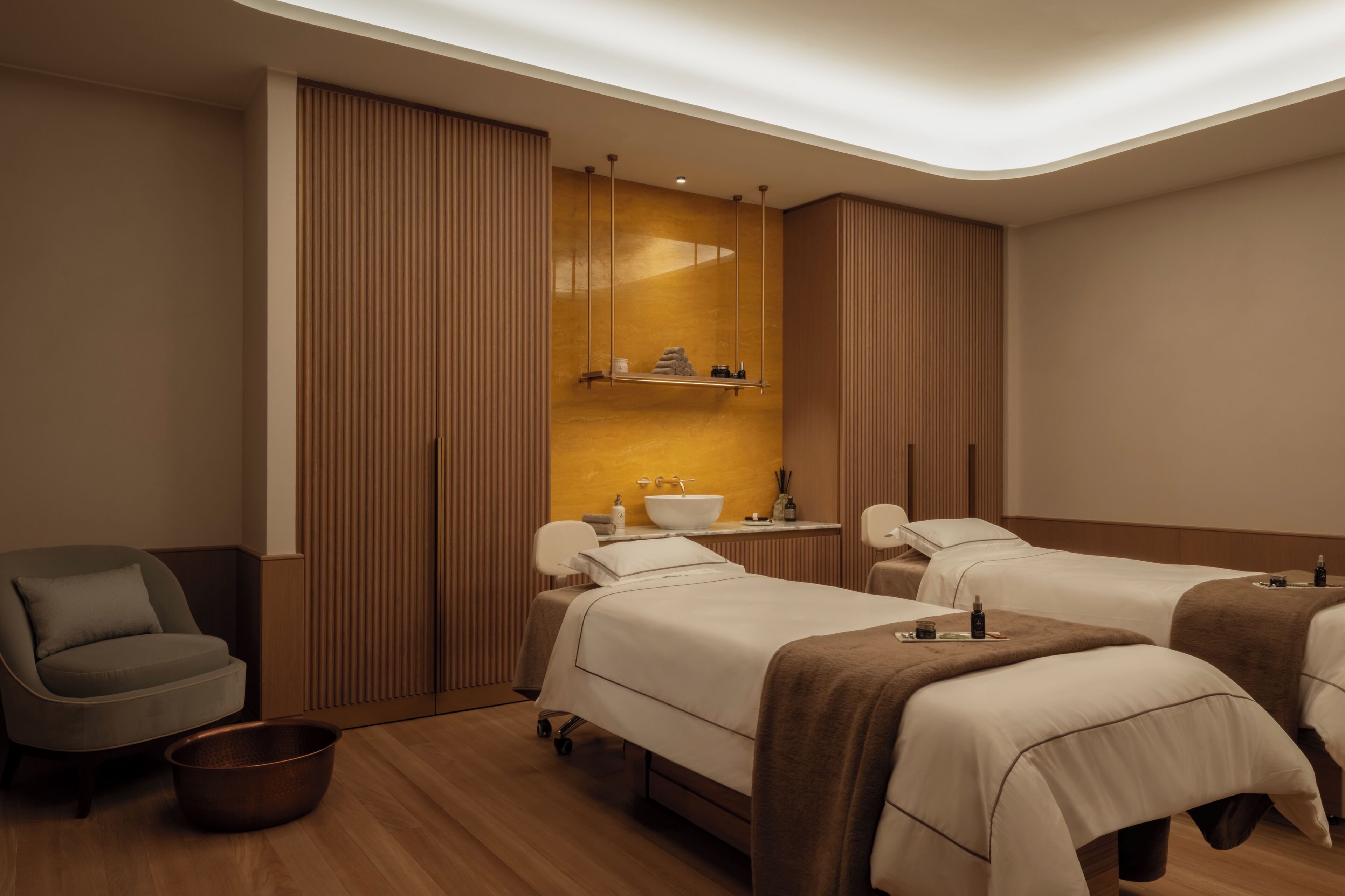 A hotel room with two beds, a chair, a small table, a sink, and a mirror.