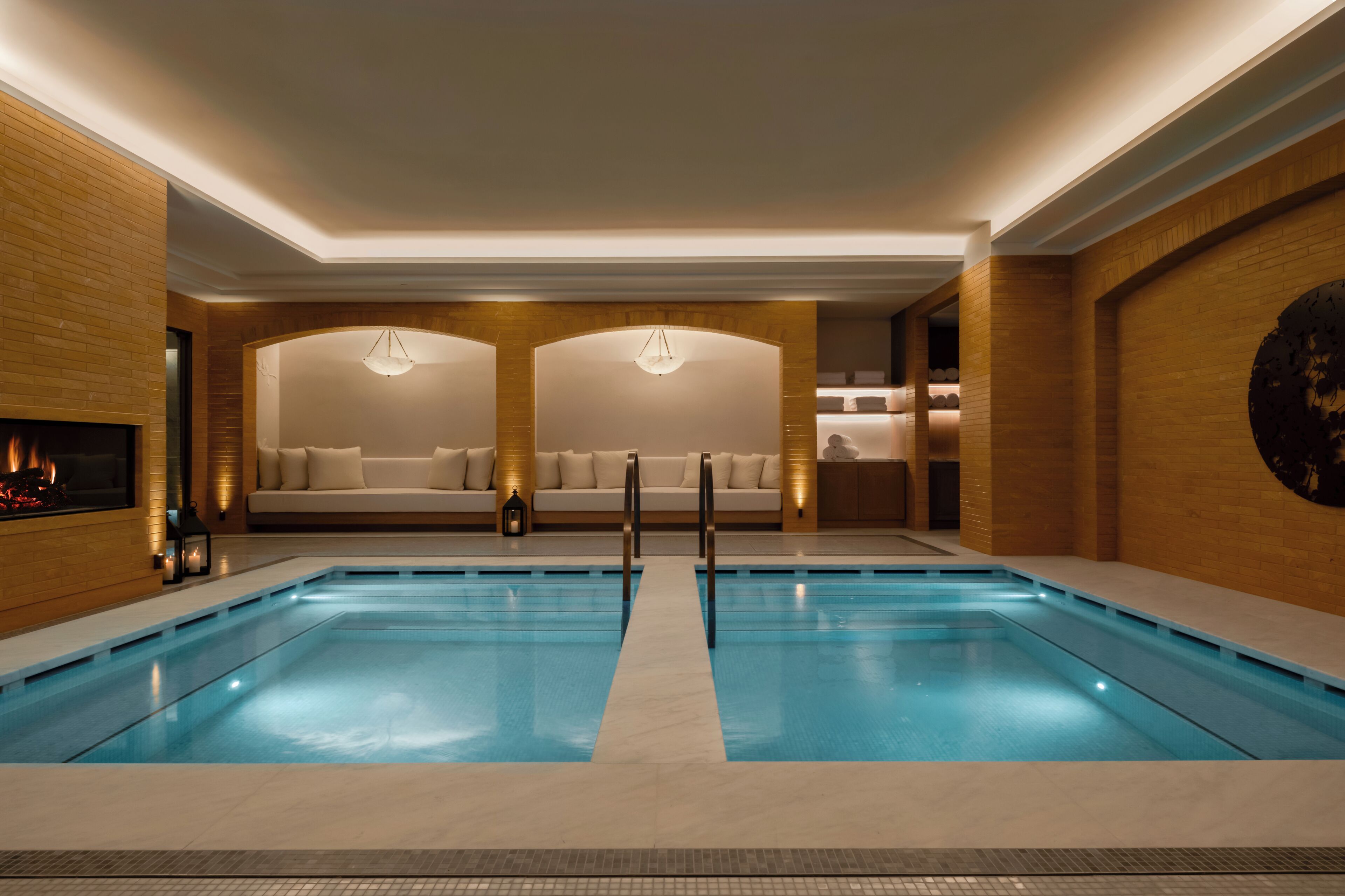 A modern indoor swimming pool with a central walkway, two sofas with cushions, and a fireplace.