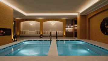 A modern indoor swimming pool with a central walkway, two sofas with cushions, and a fireplace.