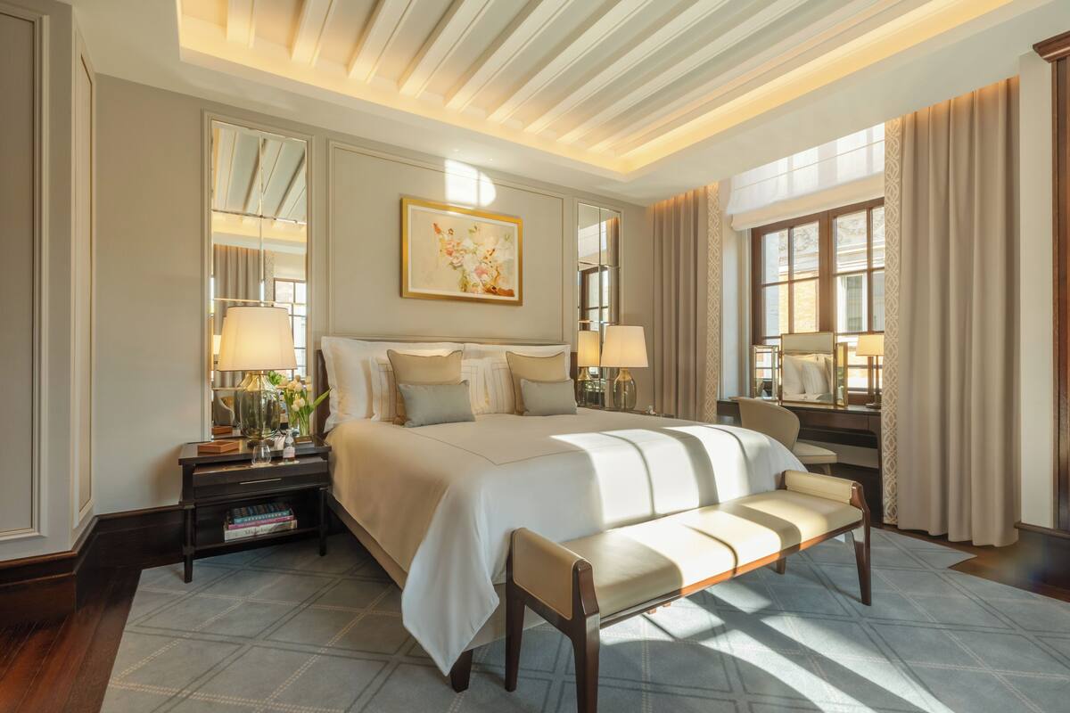 A well-lit hotel room with a large bed, bedside tables, a bench, and a large window with curtains.
