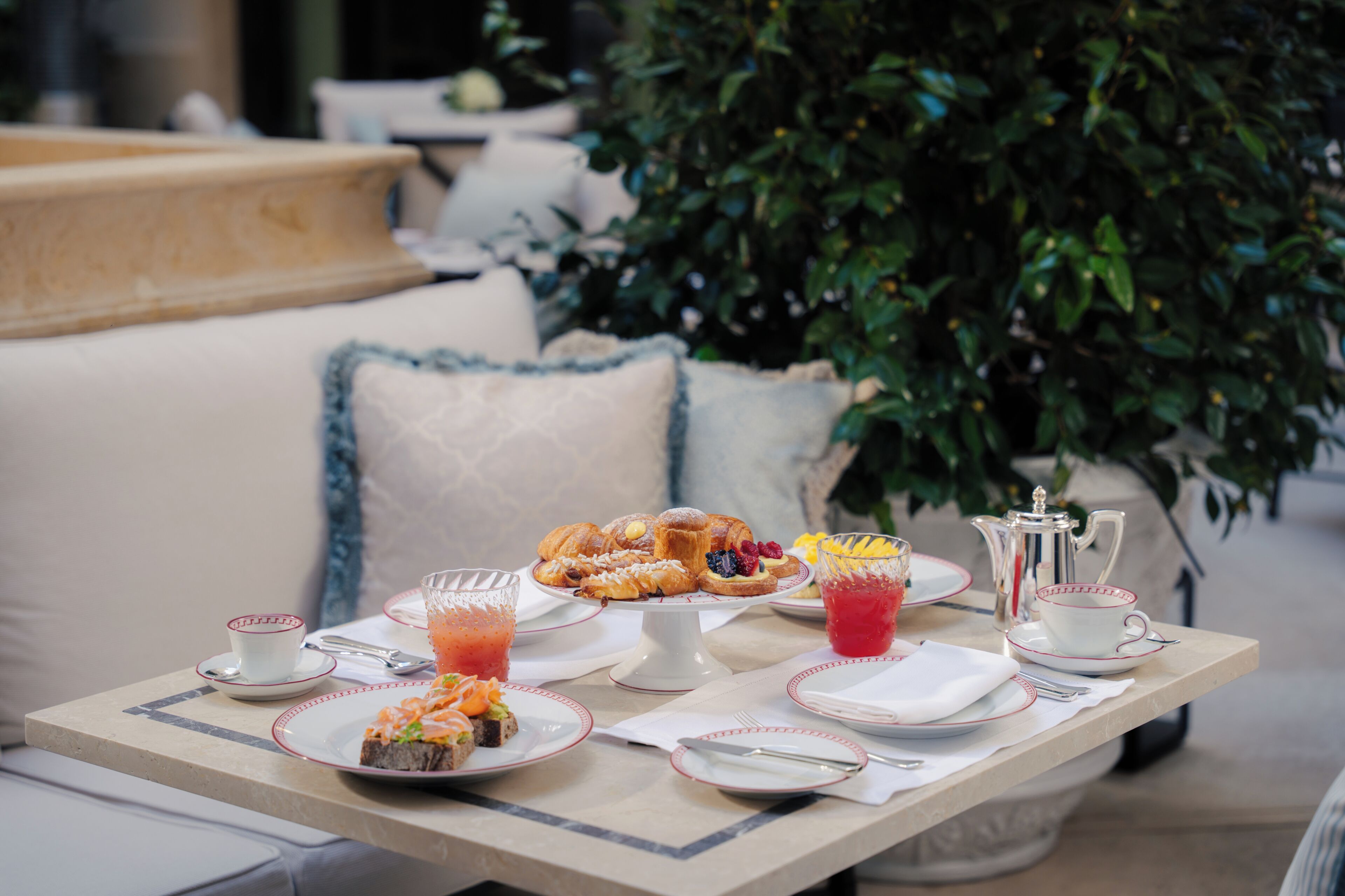 A table set with breakfast items, a teapot, and cups, with a sofa and a plant in the background.