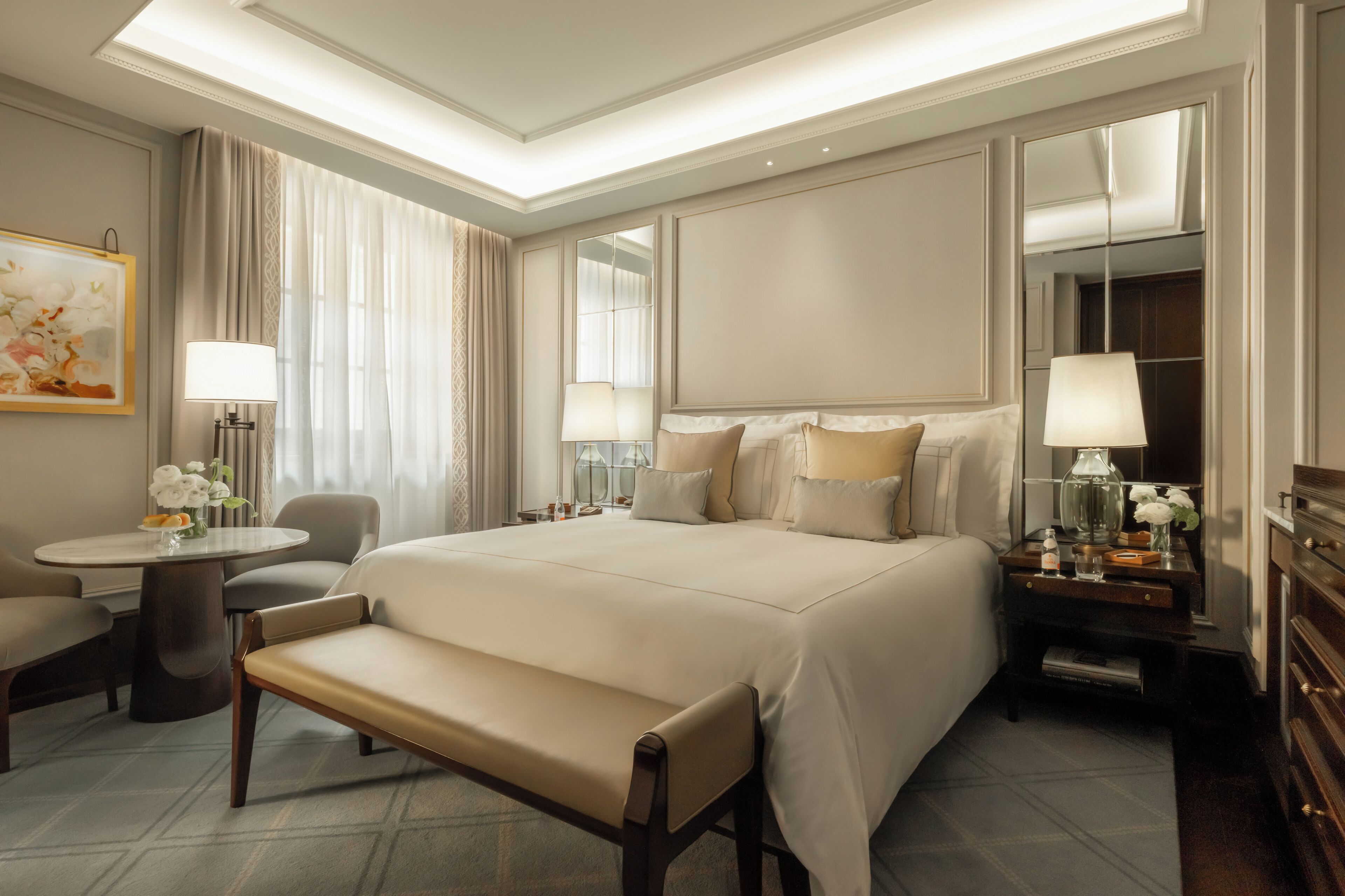 A hotel room with a large bed, a sitting area with a bench, a round table with a lamp, and a mirror on the wall.