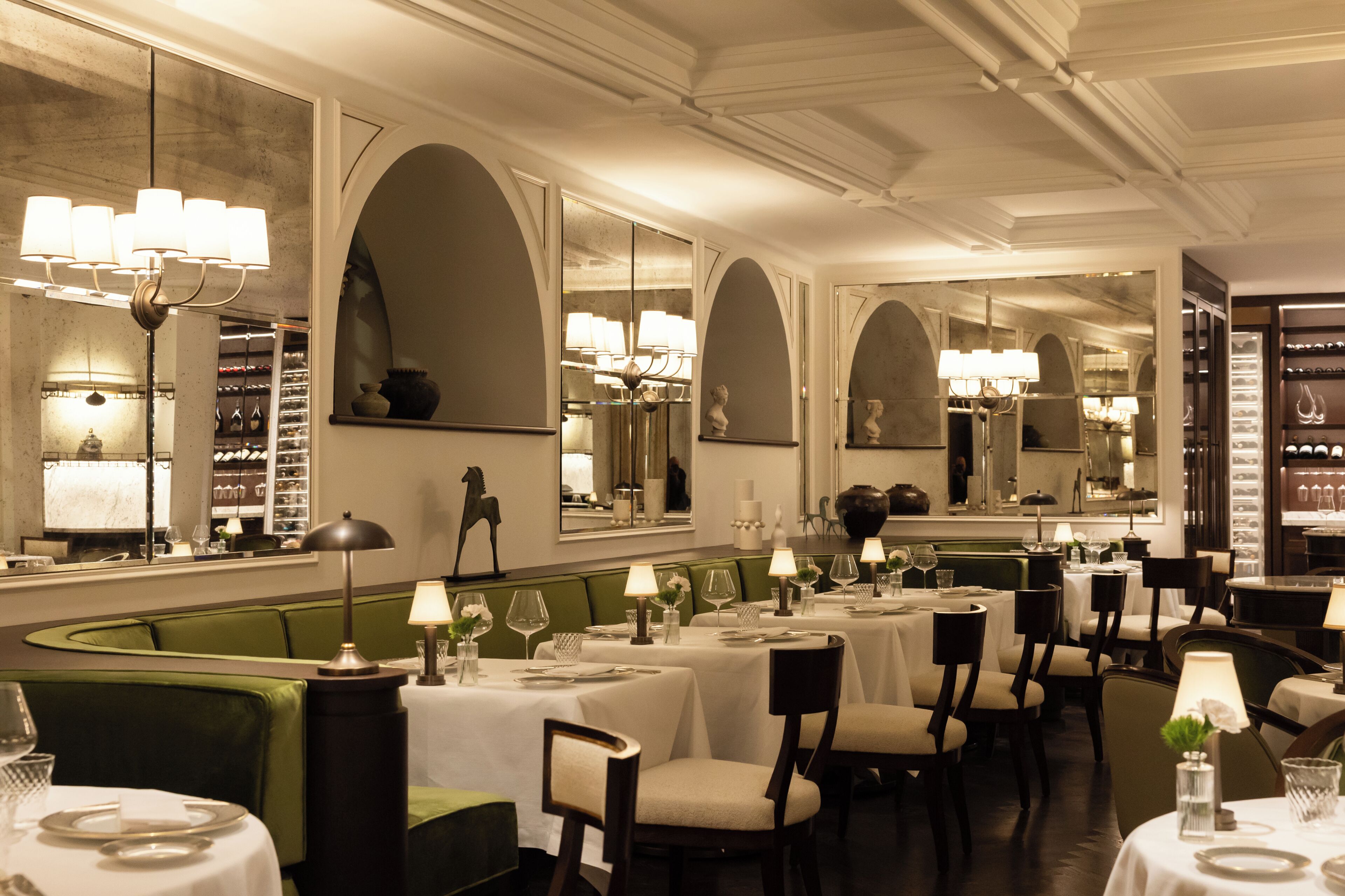 A restaurant with multiple tables, green cushioned seating, and chandeliers.