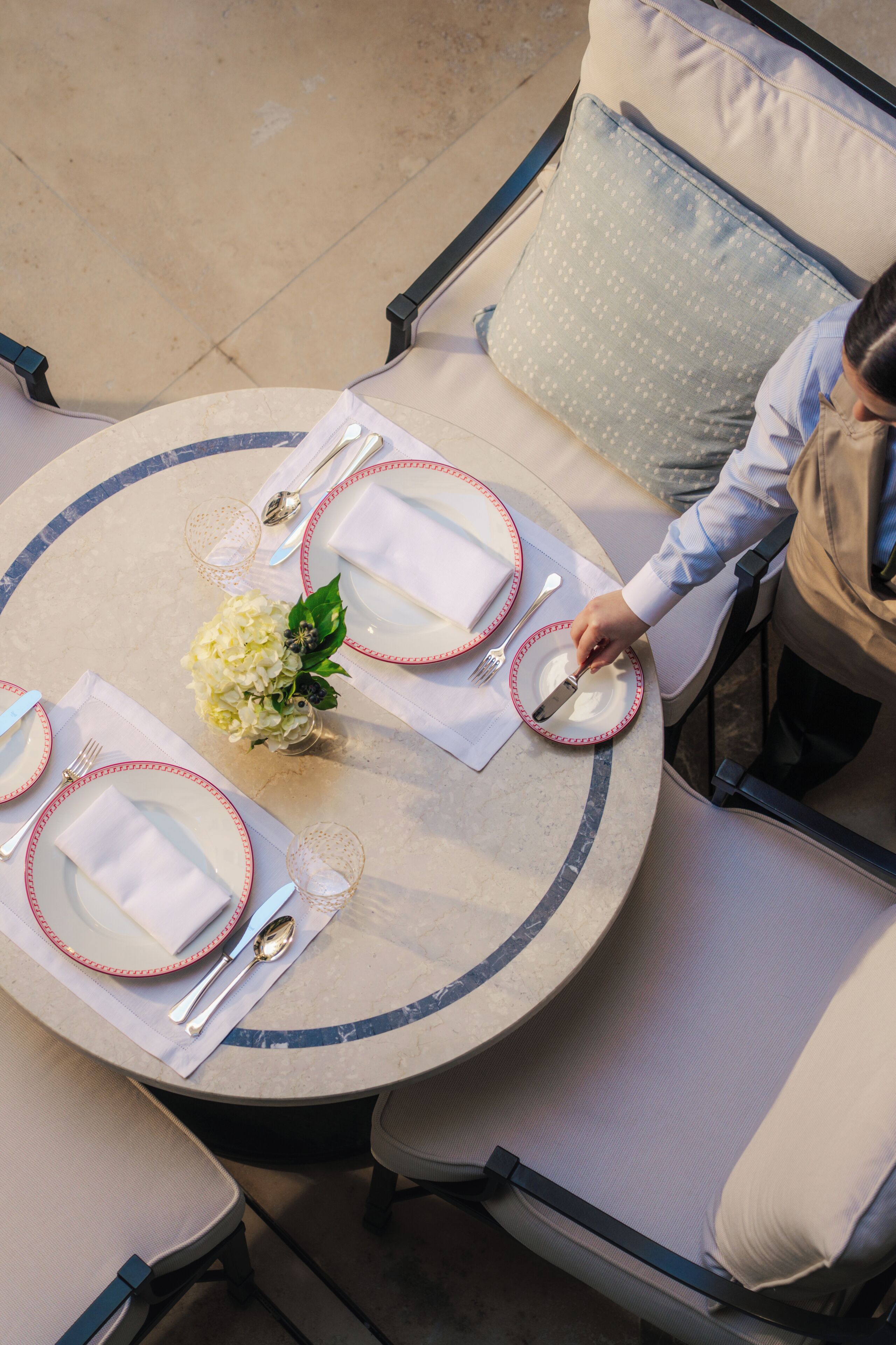 A round dining table with white plates, red rimmed cutlery, and a vase of white flowers.