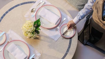 A round dining table with white plates, red rimmed cutlery, and a vase of white flowers.