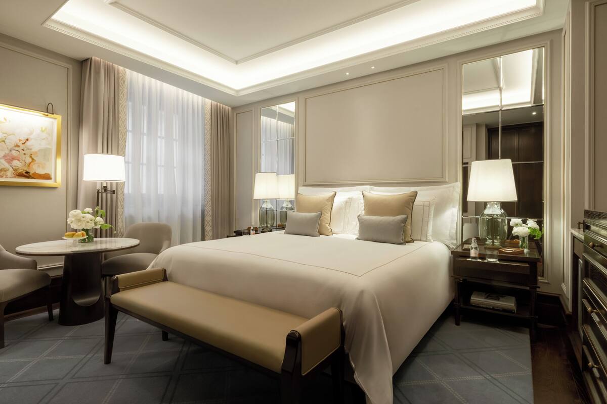 A hotel room with a large bed, a sitting area with a bench, a small table with a lamp, and a mirror.