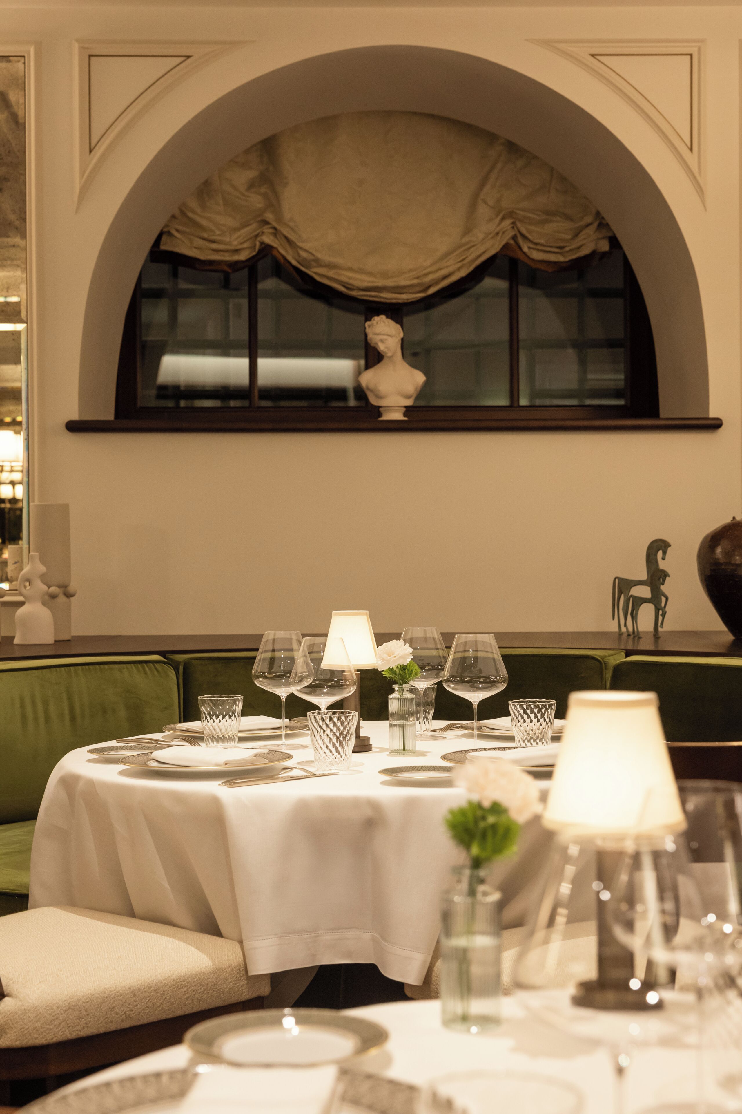 A dining table set with plates, glasses, and a vase of flowers, under a large arched window with a statue.