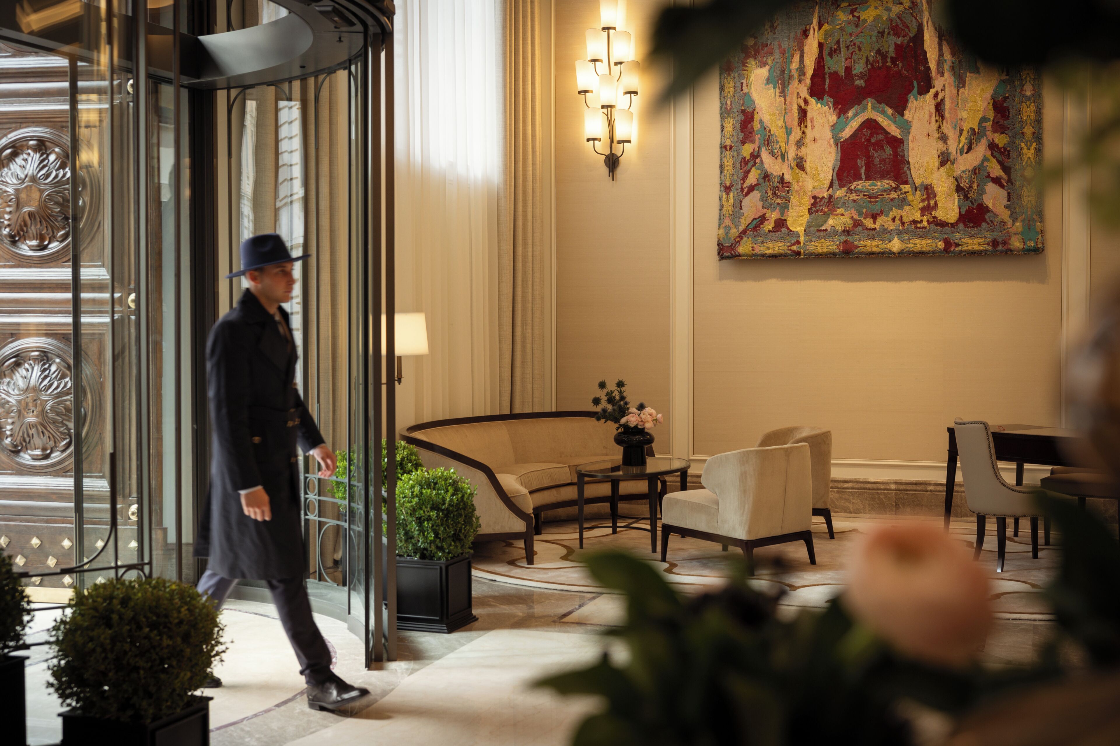 A man in a suit walking through a revolving door into a hotel lobby with a curved sofa, a table with flowers, and a large painting on the wall.