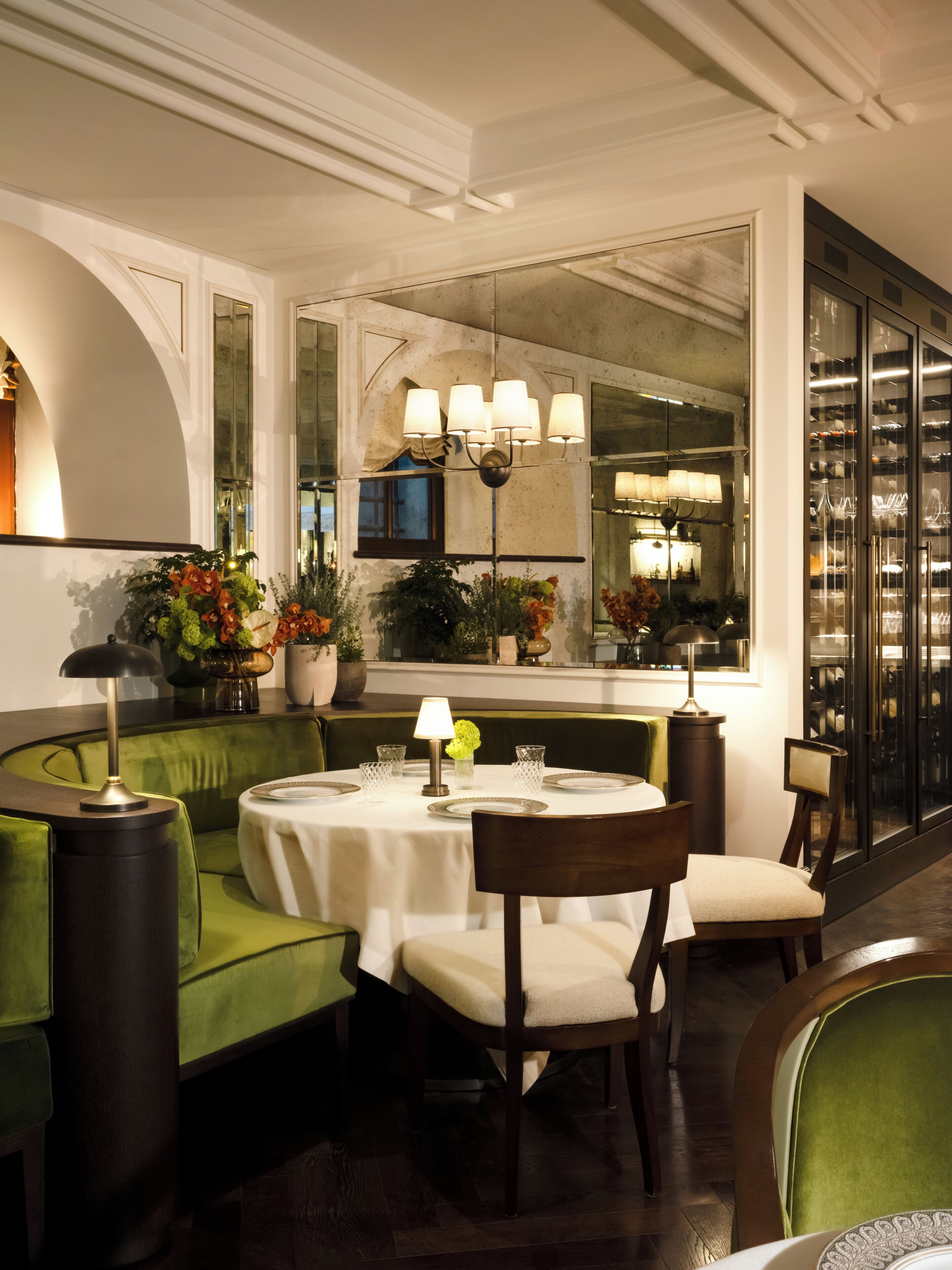 A dining area with a round table, chairs, a green sofa, a mirror, and a view of the kitchen through a window.