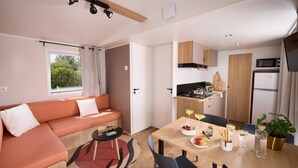 En kompakt opholdsstue med en sofa, et lille spisebord med stole, et tekøkken med mikroovn og et vindue med gardiner.
