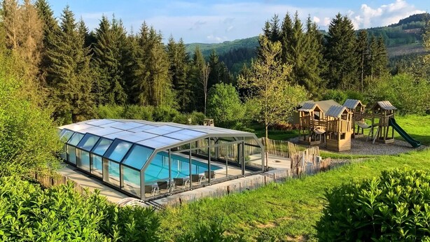 Una piscina con cerramiento de vidrio, rodeada por una terraza de madera y un parque infantil.