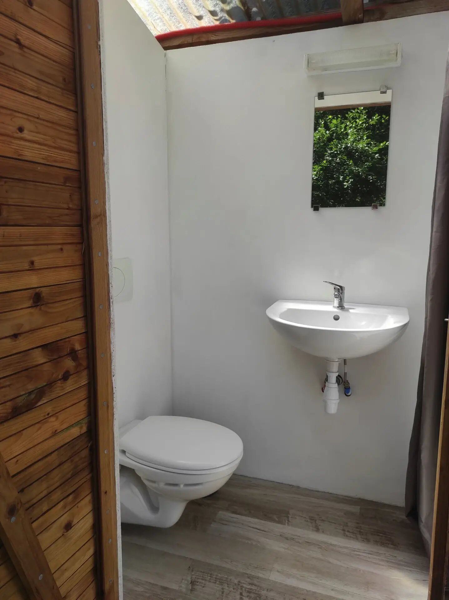 Een kleine, praktische badkamer met een wit toilet, een witte wastafel en een klein raam met uitzicht op groen.