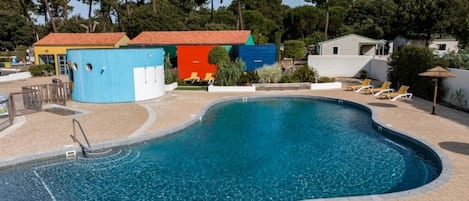 Una piscina con borde curvo, rodeada por una solana con baldosas. Hay sillas de descanso y un pequeño edificio con techo rojo.