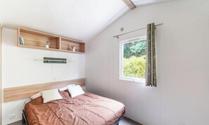 Quarto com cama, janela com cortinas e prateleira com livros e planta.