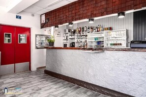 Eine Bar mit Regalen voller Flaschen und Gläsern, einer roten Tür und einer weißen, strukturierten Wand.