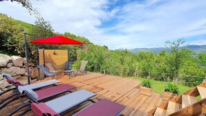 Une terrasse en bois, dotée de chaises longues et d’un parasol rouge, offre une vue imprenable sur un paysage verdoyant et luxuriant, avec des montagnes qui se dessinent à l’horizon.