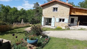Une maison en pierre avec une terrasse en bois, un jardin fleuri et une allée gravillonnée.