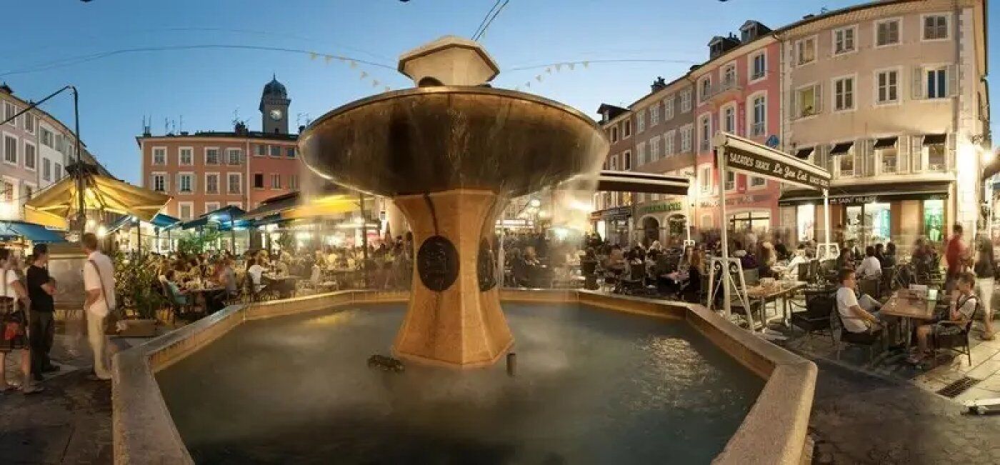 Ett offentligt torg med en central fontän, omgivet av kaféer och butiker.