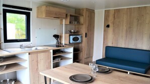 A compact kitchenette with a microwave, sink, and wooden cabinets. There is a dining table with plates and glasses, and a blue sofa. - ZEcamping 4* familial piscine couverte jacuzzi sauna locatif TV 24m2 (Caro)