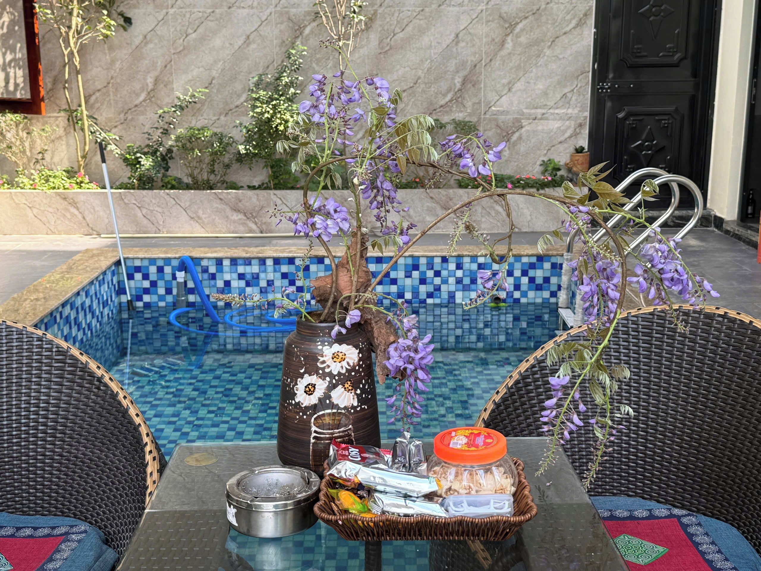 A poolside setting with a table, wicker chairs, a vase with flowers, and a tiled pool.