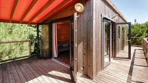A wooden deck with a red canopy, a sliding glass door leading to a room with a sofa, and a view of greenery.