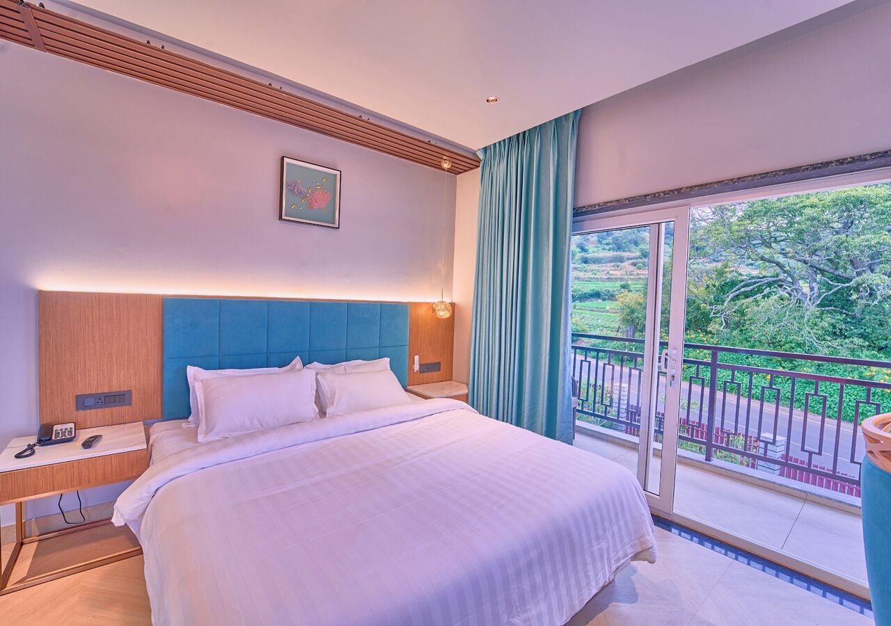 Une chambre d’hôtel avec un grand lit, une table de chevet avec un téléphone, une lampe fixée au mur et une vue sur un balcon agrémenté de verdure.
