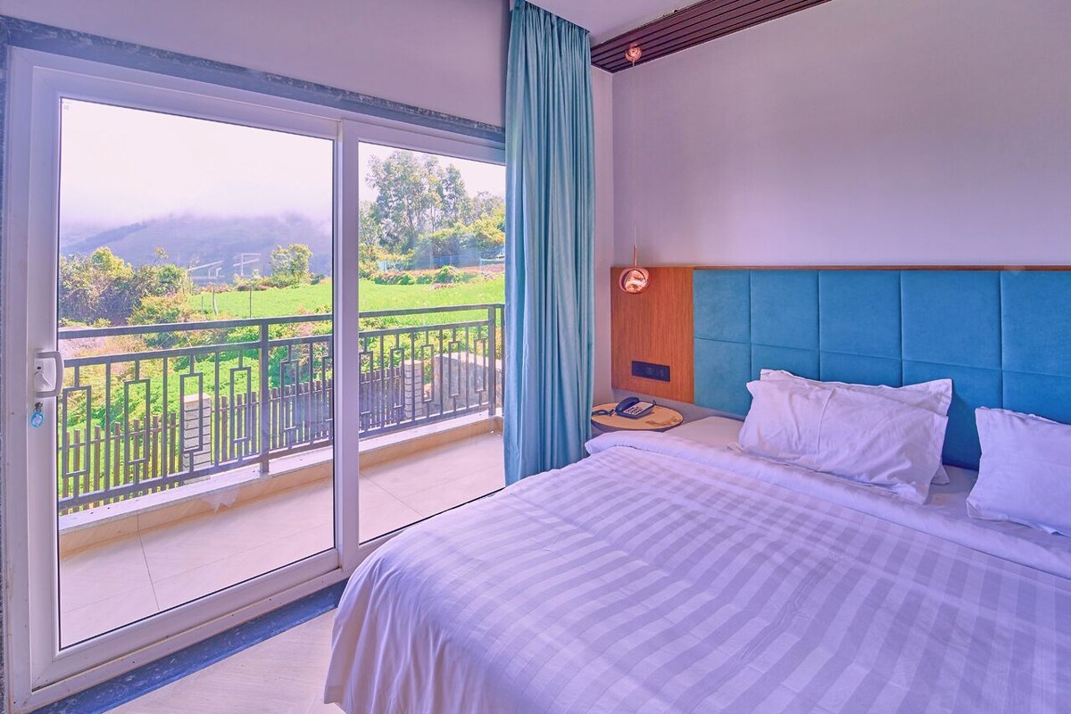 A hotel room with a large bed, a wooden headboard, a nightstand, and a view of a green landscape through a sliding glass door.