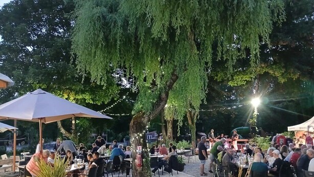 Un espace de restauration en plein air, avec des tables et des chaises disposées sous un grand arbre, entouré de verdure et illuminé de guirlandes lumineuses.