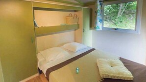 Un lit simple avec une tête de lit, une petite étagère au-dessus et une fenêtre donnant sur un paysage verdoyant.