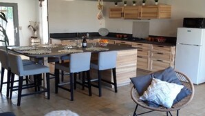 A modern kitchen with a central island, bar stools, hanging light fixtures, wooden cabinets, and a refrigerator. - ZEcamping piscine chauffée dog friendly locatif LV BBQ 90m2 6 pers (Vendoire)