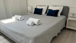 A neatly made bed with a grey bedspread and two rolled white towels, two dark blue pillows, and a white nightstand with a lamp. - In Barcelos View Lodge (Barcelos)
