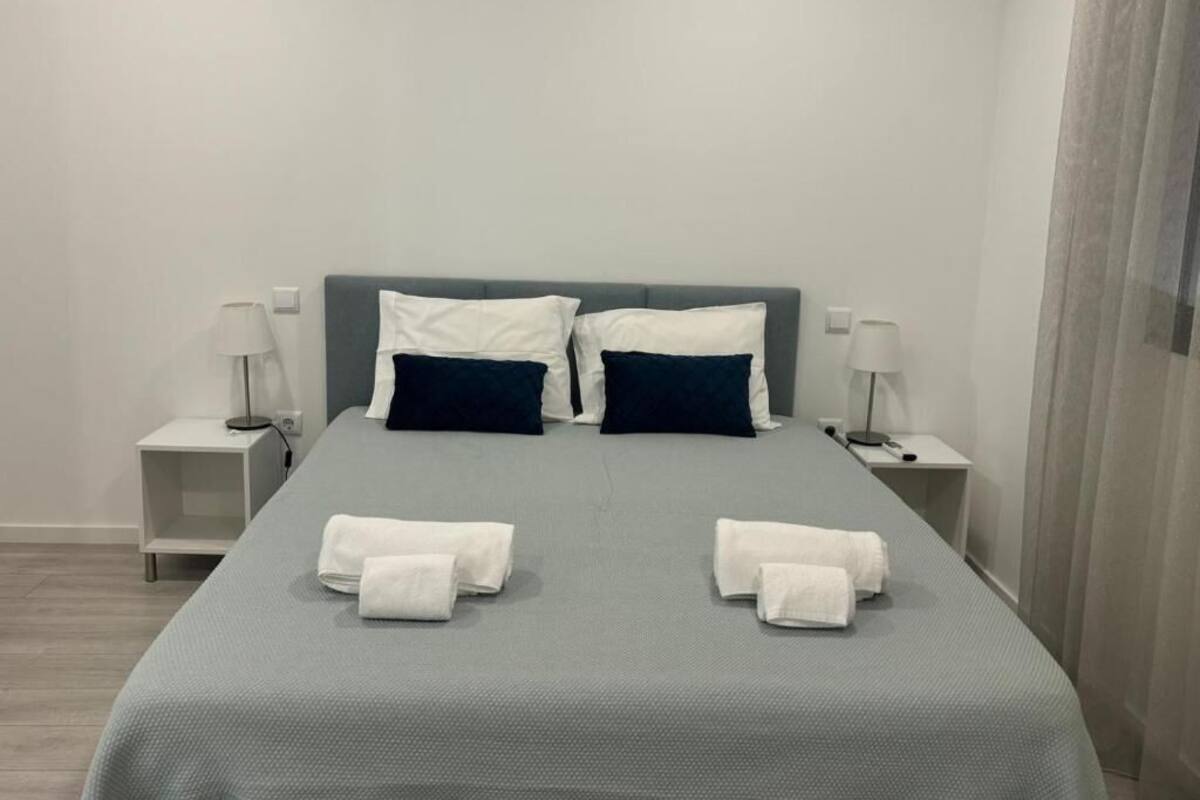 A neatly made bed with white and navy pillows, a grey bedspread, and two white bedside tables with lamps.