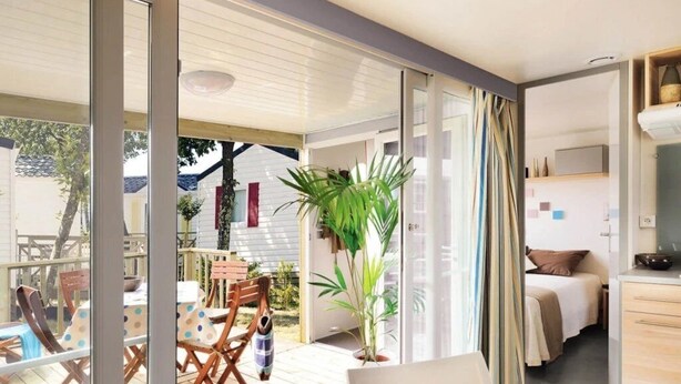 Une chambre moderne dotée d’une porte-fenêtre coulissante donnant sur une terrasse en bois, d’un coin repas avec une table et des chaises, et d’un espace cuisine avec un lit et une table de chevet.