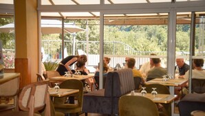 Ein Essbereich im Freien mit Tischen und Stühlen, ein Kellner bedient Gäste, und im Hintergrund ist viel Grün zu sehen.