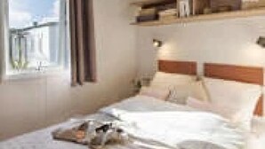 A bedroom with a bed, a window with curtains, a shelf with items, and a plant. - ZEcamping 3* pataugeoire piscine dog friendly locatif TV clim 26m2 (Les Assions)