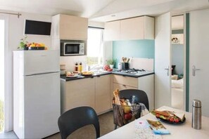A compact kitchen with a refrigerator, microwave, and stove, a dining table with chairs, and a small window. - ZEcamping 3 étoiles pataugeoire locatif TV clim 31m2 (Les Assions)