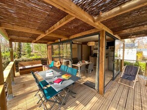 Un patio exterior con terraza de madera, una mesa de comedor con sillas, un sofá y una pequeña zona de cocina con refrigerador y fregadero.