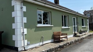 A single-story house with a green exterior, white trim, and a chimney. There is a bench outside and a small garden area with potted plants. - Christinas Cottage - Family- Garden-wifi-sleeps 6 (Downpatrick)