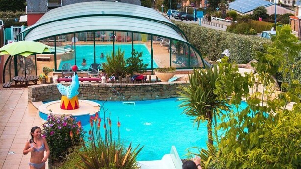 Une piscine avec un toboggan, entourée de verdure et dotée d’un espace abrité avec des sièges.