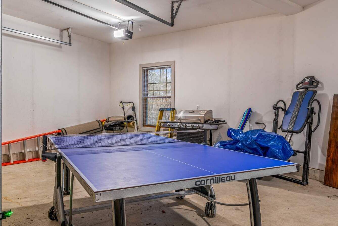 A ping pong table with a blue playing surface, a projector, and exercise equipment in a room with white walls and a window.