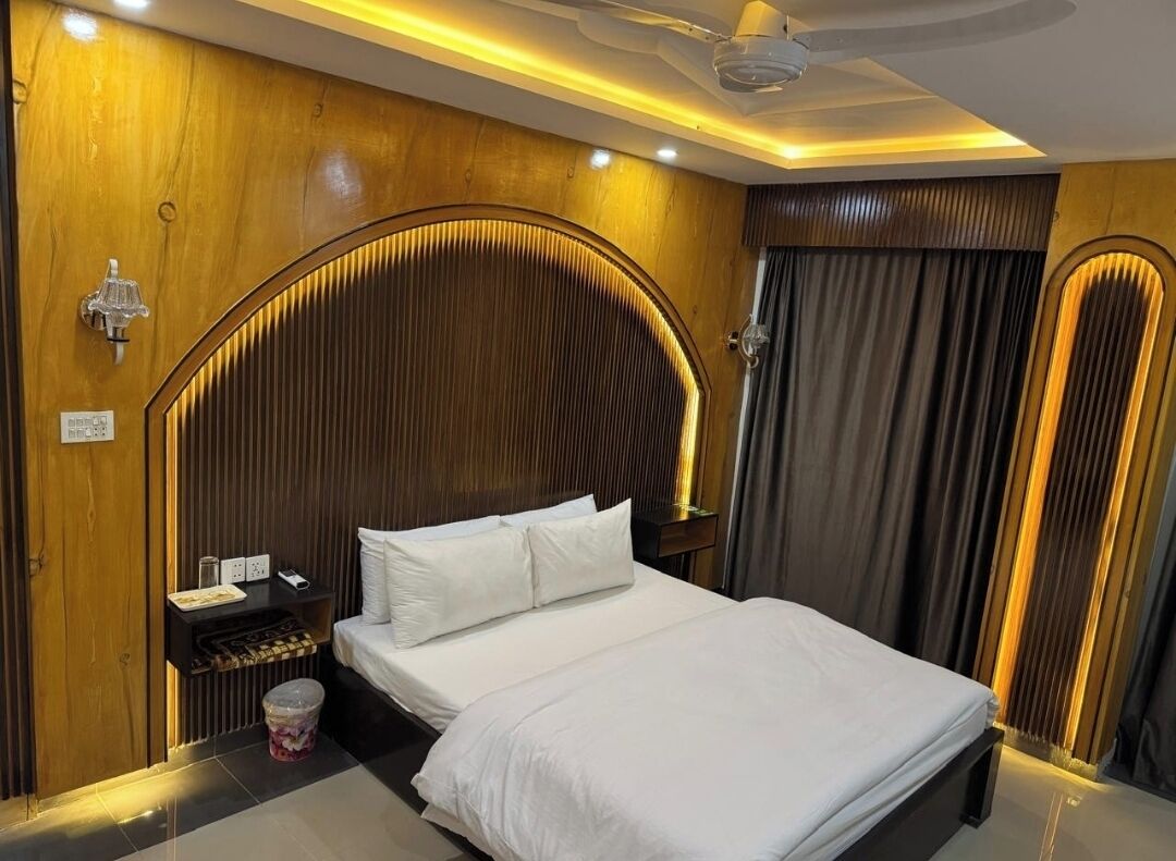 Quarto de hotel com uma cama grande, painéis de madeira, uma mesa de cabeceira com abajur e um ventilador de teto.