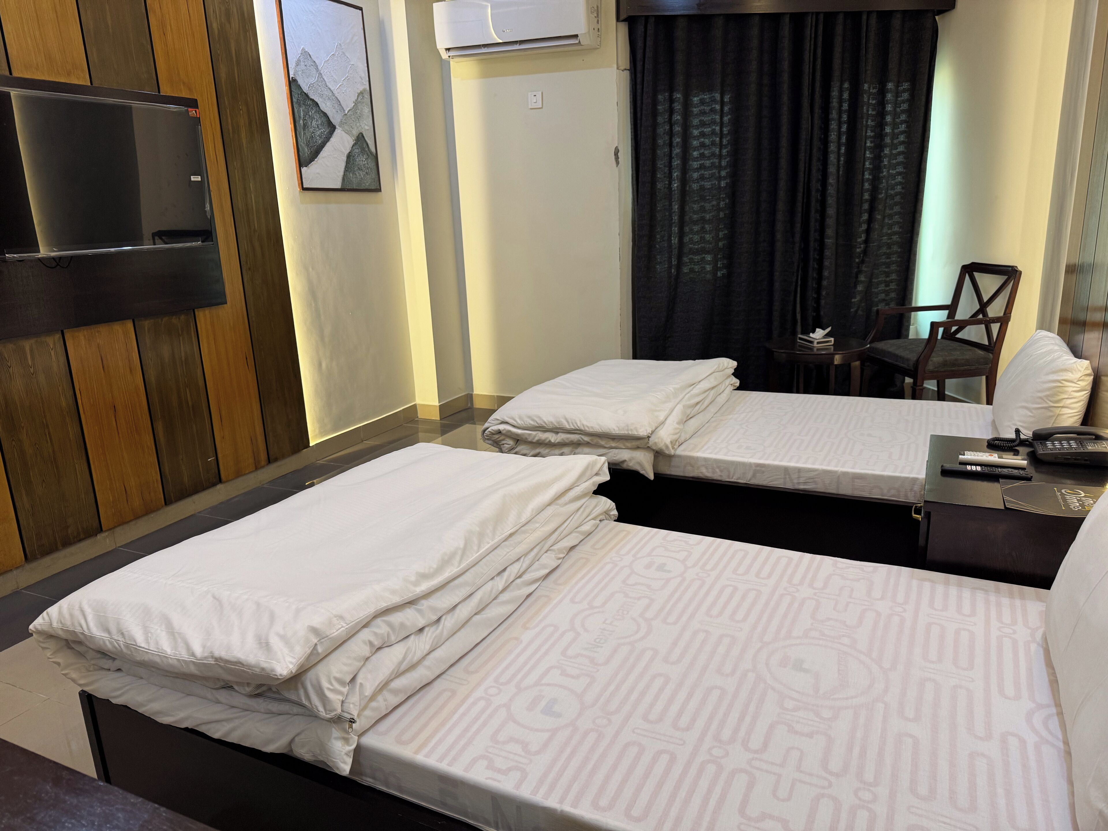 Une chambre d’hôtel avec deux lits, un téléviseur à écran plat fixé au mur, un bureau avec un téléphone et une télécommande, et une fenêtre avec des rideaux.