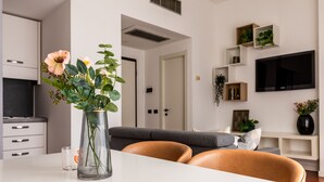 A modern living room with a white sofa, a glass vase with flowers, a flat-screen TV, and built-in shelving with plants. - Garibaldi - Modern 1 BR in Brera by Rentopolis (Milan)