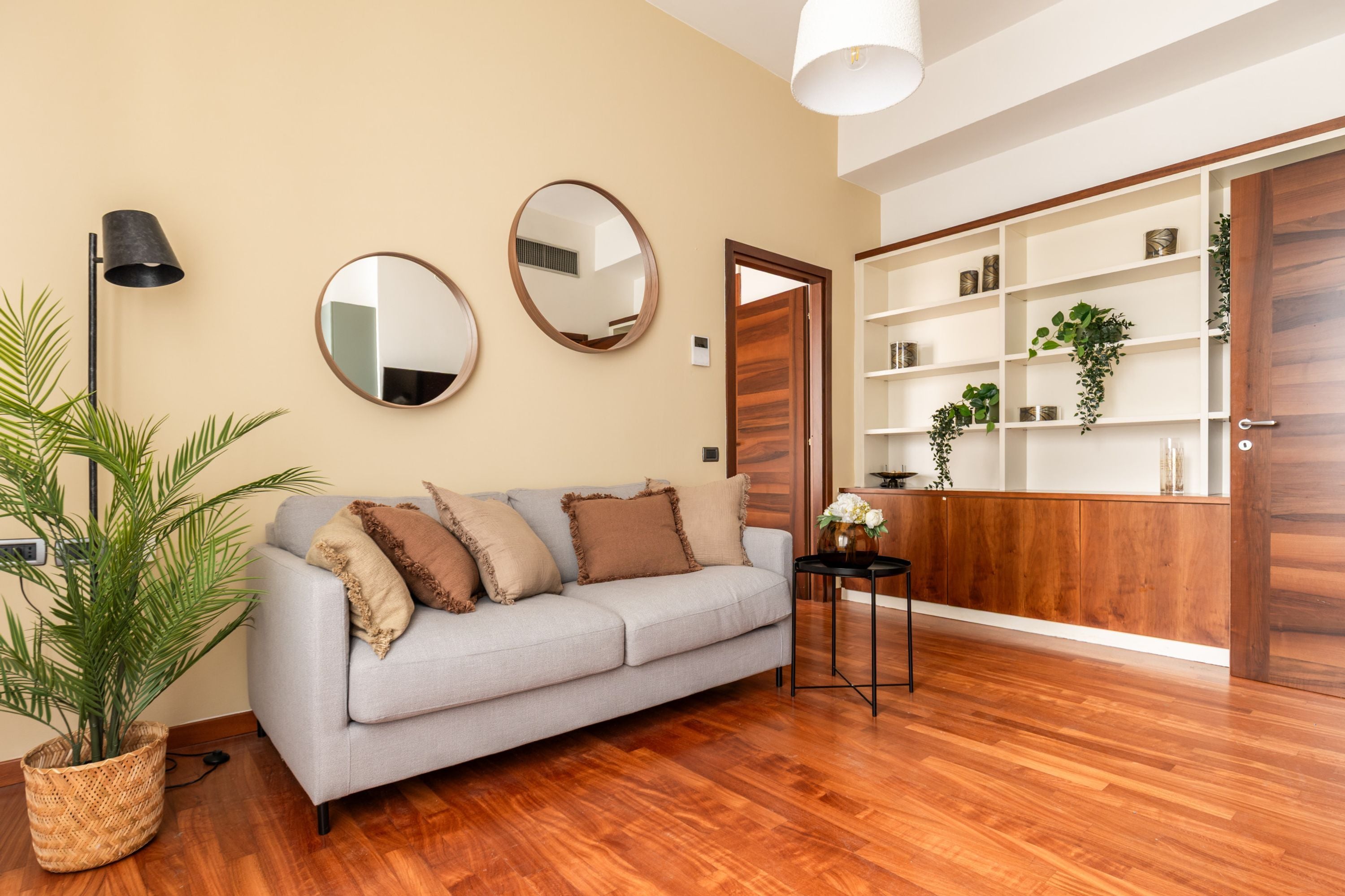 Una sala de estar con un sofá gris, dos espejos redondos, una lámpara de pie, una planta en maceta y un mueble de madera con estantes.