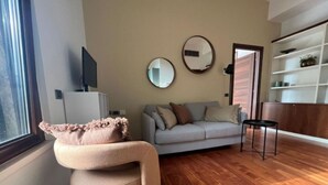 Una sala de estar con sofá, una mesita, una silla y televisor sobre un mueble bajo. Se observan dos espejos en la pared y una estantería con objetos.