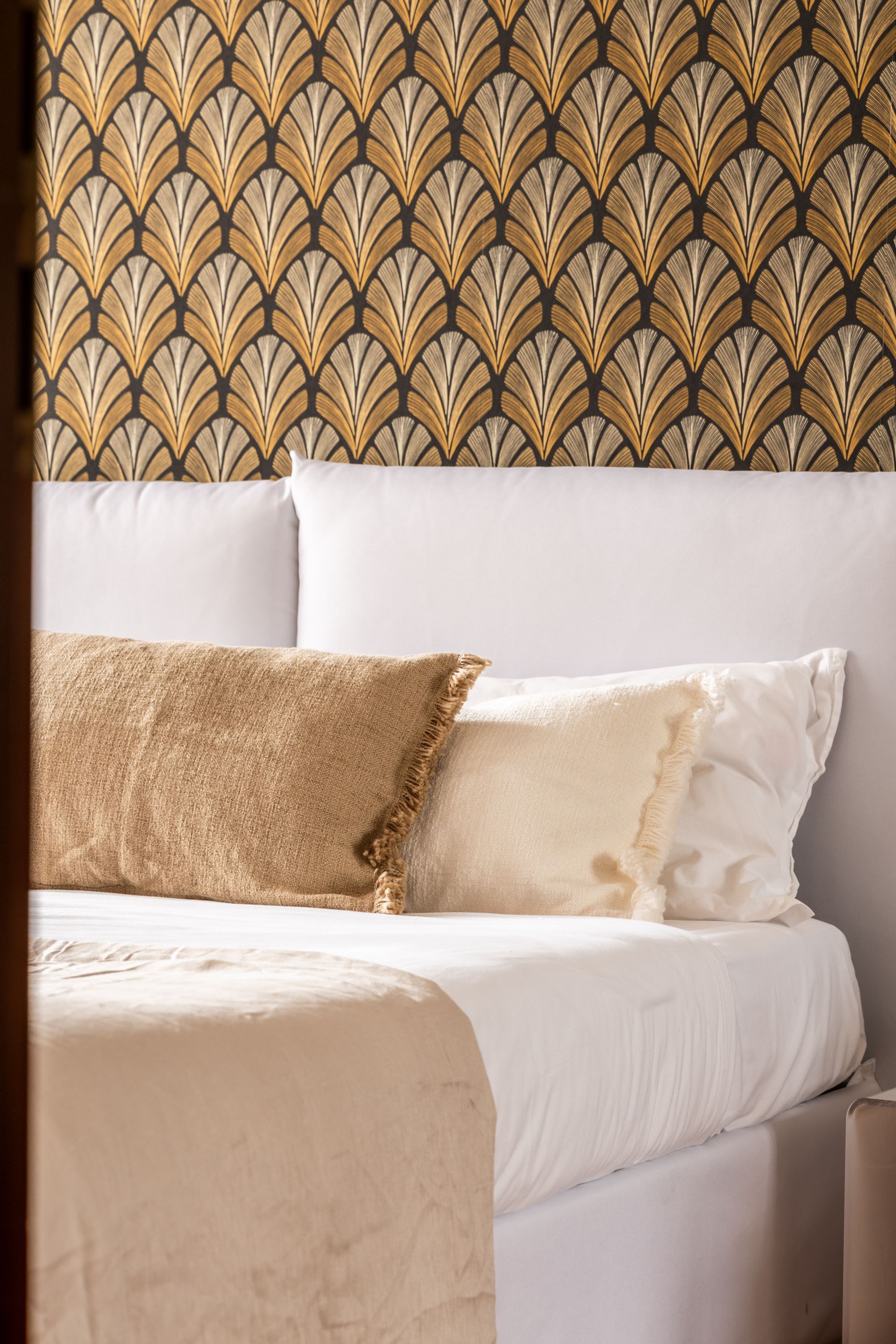Una cama con ropa blanca y dos almohadas, una de tela texturizada y la otra lisa, frente a una pared con papel tapiz estampado.